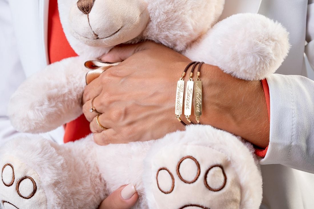 Mãos da Dra. Priscilla Aquino abraçando um urso de pelúcia, com pulseiras gravadas com os nomes Priscilla, Lucas e Matheus
